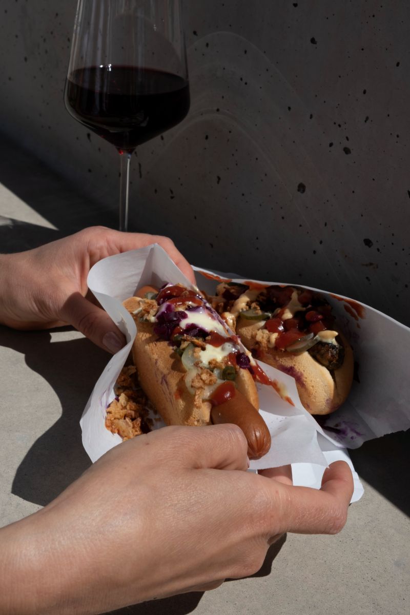 Eine Person isst einen Hot Dog und trinkt dazu Rotwein.