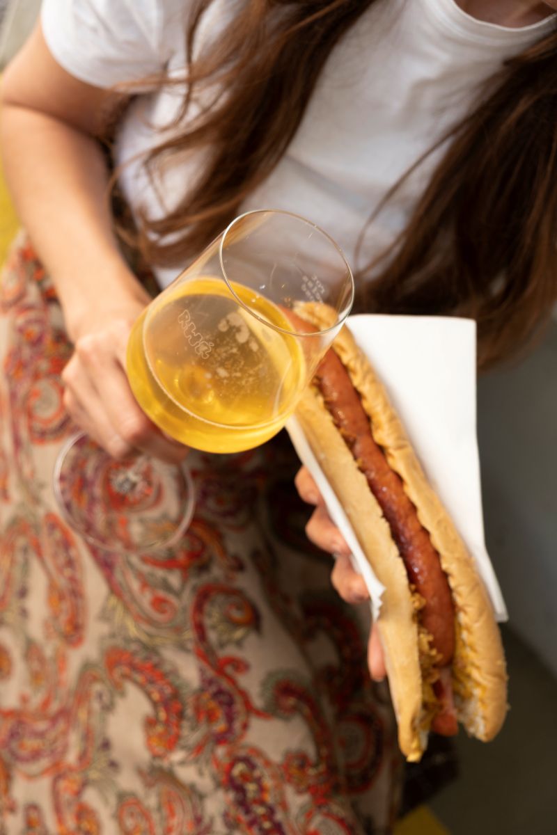 Eine junge Frau hält eine Bosna und ein Glas Pet Nat.