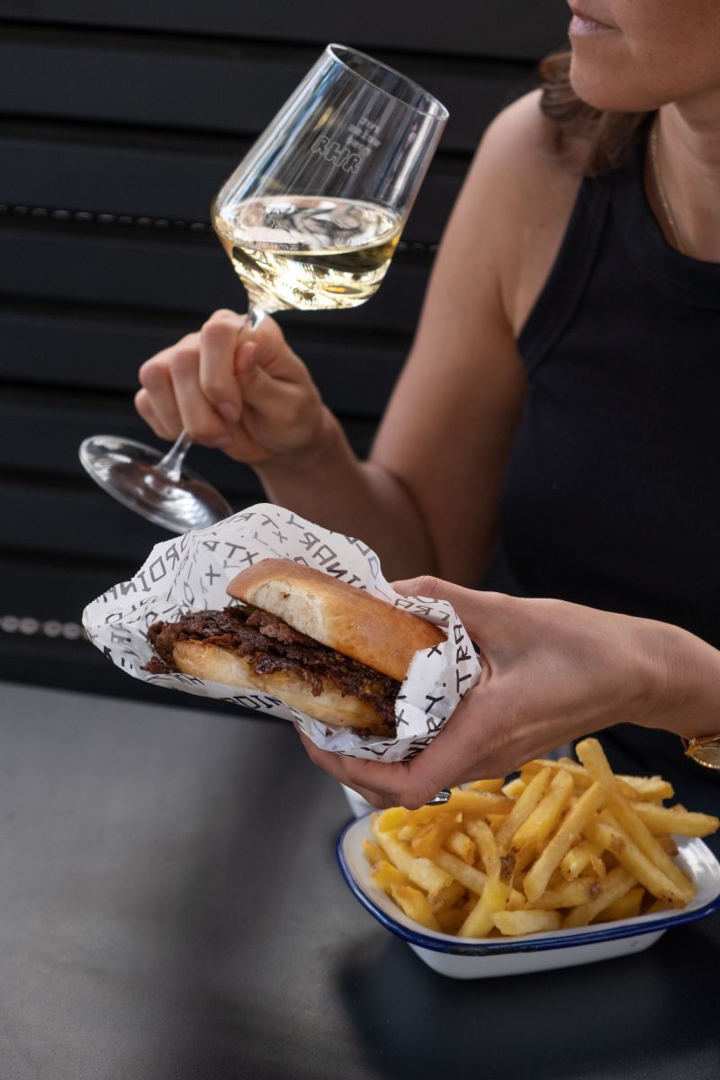 Das Bild zeigt eine junge Frau mit Burger und Weißwein.